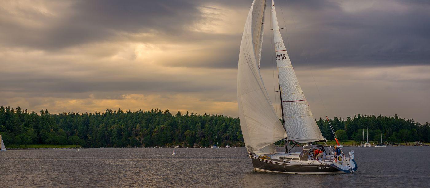 A sailboat under full sail on coastal water beneath a heavy, broken sky
